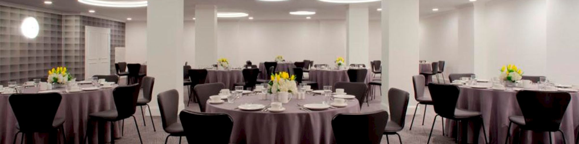 The image shows a banquet hall with round tables covered in gray tablecloths, neatly arranged chairs, and floral centerpieces.