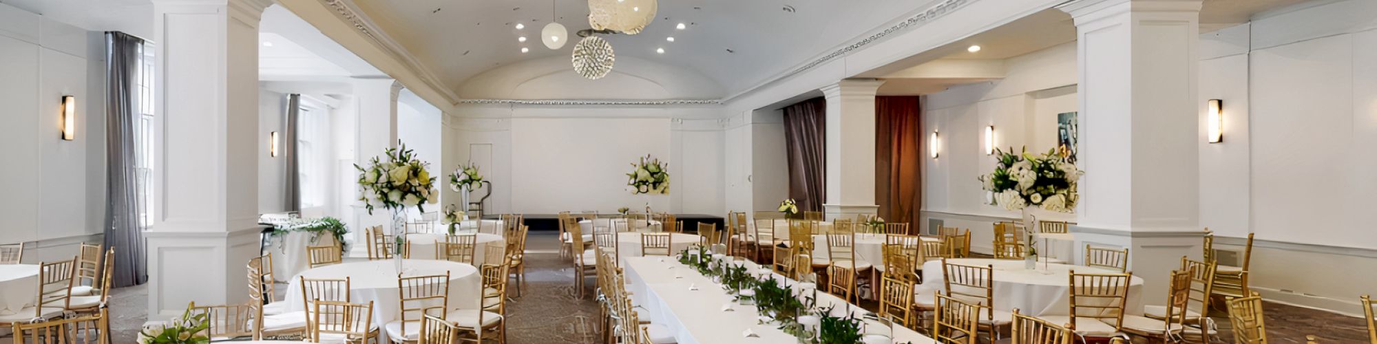 The image shows a decorated banquet hall with round and rectangular tables, gold chairs, floral arrangements, and overhead spherical lights.