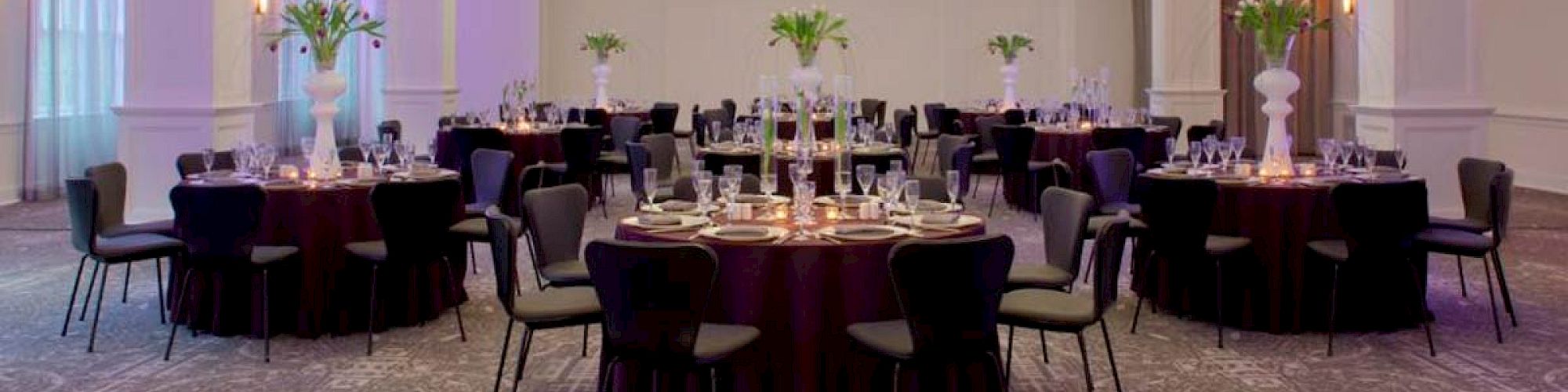 A well-decorated banquet hall with round tables, chairs, floral centerpieces, and elegant lighting fixtures hanging from the ceiling.