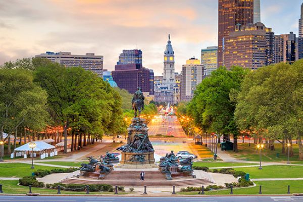 This image shows a city skyline with a prominent statue in the foreground, surrounded by trees and a park, under a colorful sky at sunset.