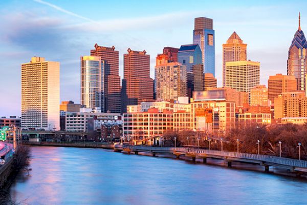 This image shows a city skyline at sunset, with tall buildings reflecting the sunlight, a river in the foreground, and a bridge crossing over the water.