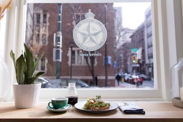 A plant, cup, and plate with food on a table by a window, showing a city street outside and a logo on the glass.