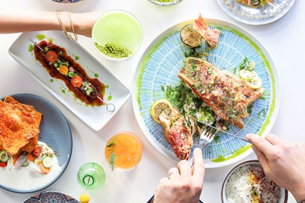 A table with various dishes: grilled fish, appetizers, rice, and colorful drinks. Two people are seen reaching for the food and drinks.