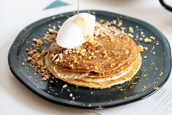 The image shows a plate of pancakes topped with whipped cream, syrup being poured, and sprinkled nuts and zest on a dark plate.