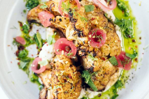 The image shows a beautifully plated dish featuring roasted cauliflower topped with pickled red onions and garnished with fresh herbs and a green sauce.