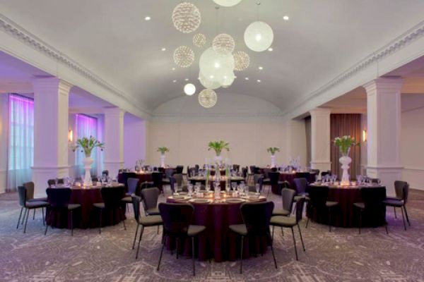 A banquet hall with round tables set for an event, decorated with white flowers and modern pendant lights hanging from the ceiling, is shown.