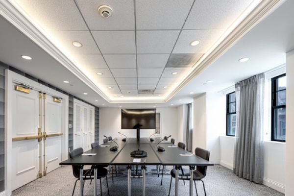 A modern conference room with a long black table, chairs, a large screen monitor, and microphones. The room has recessed lighting and windows.