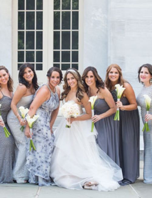 A bride in a white gown poses with her bridesmaids in gray dresses, holding bouquets, outside in front of a building with large windows.