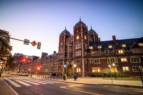 A large, historic building, likely part of a university or institution, is seen at dusk with street lights and a crosswalk in the foreground.