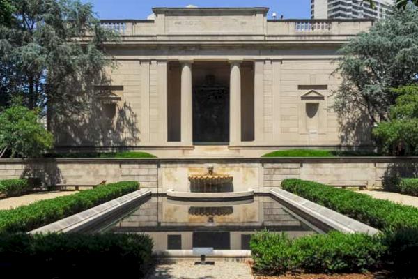 The image shows a neoclassical building with pillars at the entrance, surrounded by greenery, and a reflecting pool in front of it.