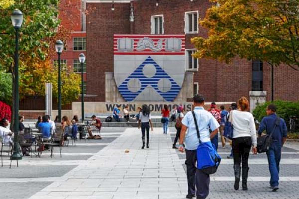 People are walking and sitting near a building with a large University of Pennsylvania logo and a sign reading 