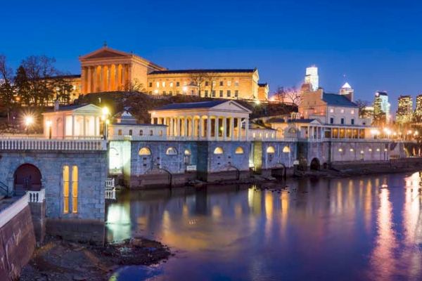 The image shows the Philadelphia Museum of Art and the Fairmount Water Works in Philadelphia, beautifully lit up at dusk with city skyscrapers in the background.