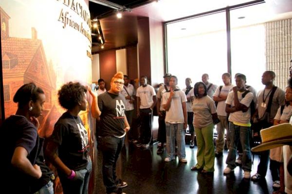 A group of people is gathered indoors, listening to a person speaking near a display. They appear to be on a tour or in an educational setting.