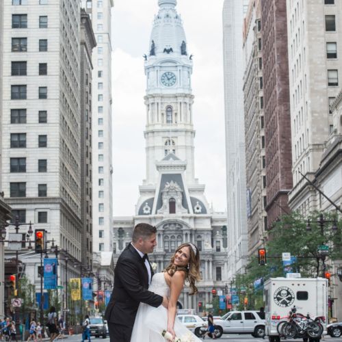 A couple in wedding attire poses in the middle of a city street with a tall, historic building in the background amidst skyscrapers and vehicles passing by.