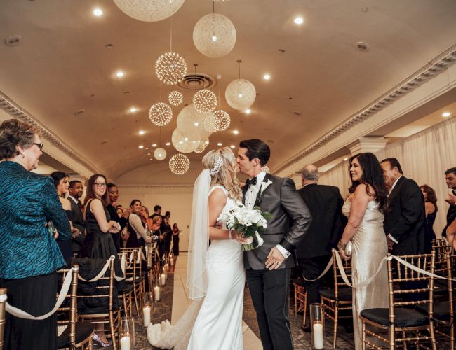 A bride and groom kiss at the end of the wedding aisle, surrounded by guests in a beautifully decorated indoor venue.