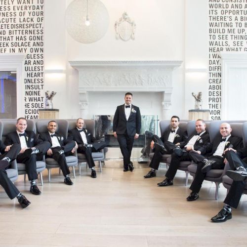 A group of men in tuxedos, seated and standing, in a stylish room with large text on the walls. They appear to be posing, possibly for an event.