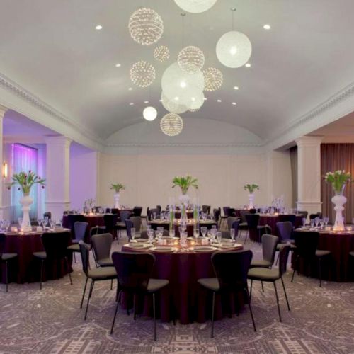 A banquet hall set up with round tables adorned with flowers, elegant lighting with hanging spherical fixtures, and comfortable chairs.