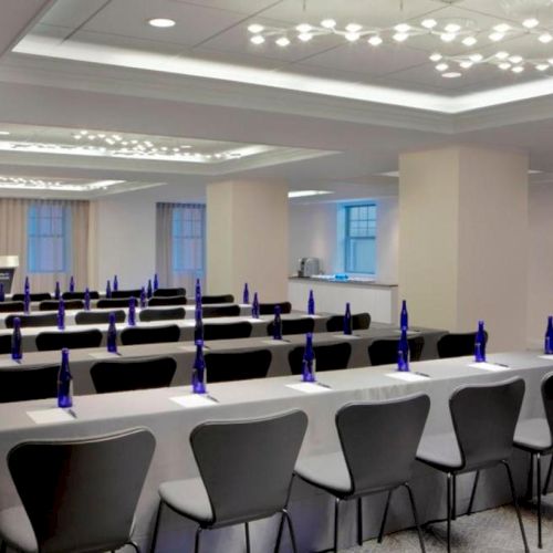 A conference room set up with rows of tables and chairs, each with blue water bottles and notepads, facing a podium and screen area at the front.