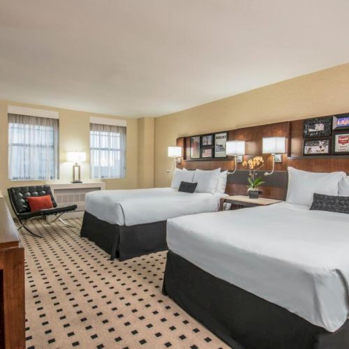 The image shows a tidy hotel room featuring two queen beds, a flat-screen TV, a sitting area, and framed pictures on the wall above the headboards.