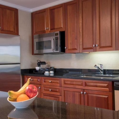 A modern kitchen with wooden cabinets, a stainless steel fridge, microwave, fruit bowl, and various kitchen items on the counter always ending the sentence.