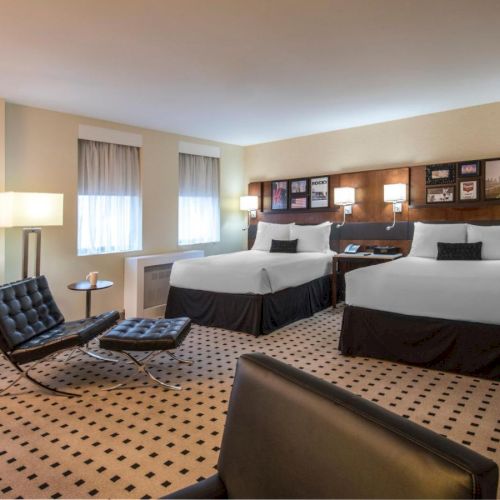 This image shows a hotel room with two double beds, modern furniture, two lounge chairs, a side table with a lamp, and a patterned carpet.