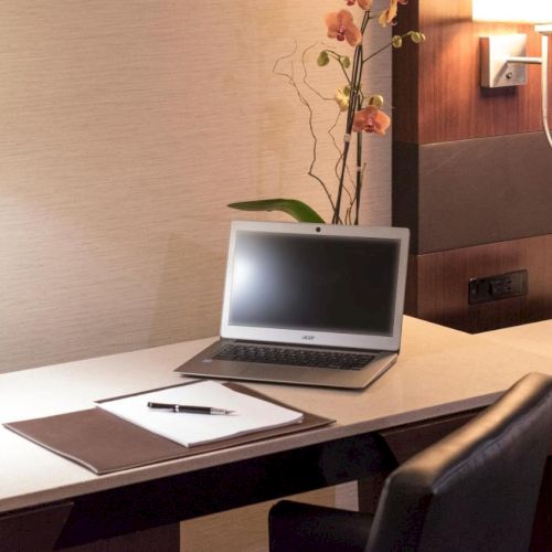 A tidy workspace with a laptop, notepad, pen, desk lamp, flowers, and a landline phone on a sleek desk with a leather chair under soft lighting.