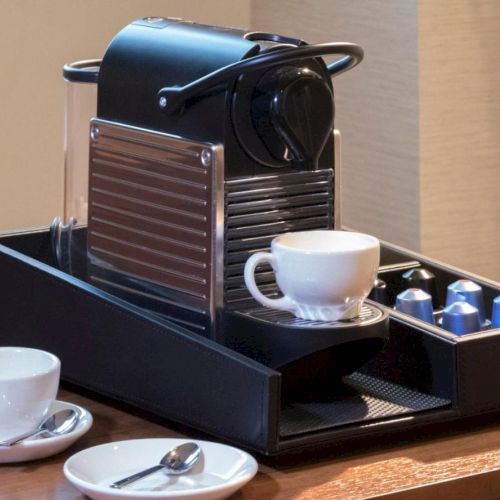 A coffee machine with a cup, surrounded by coffee pods, two empty cups, and saucers with spoons on a wooden table.