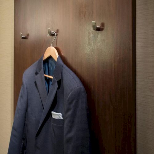 A dark blue suit jacket hangs on a wooden panel with three coat hooks in a dimly lit room.