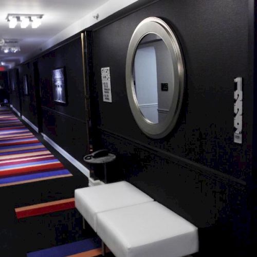 A modern hallway featuring a white bench, round mirror, black walls, and a colorful carpet with red, blue, and orange stripes.