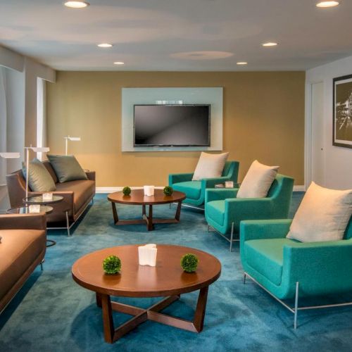 A modern living room with teal armchairs, brown sofas, a flat-screen TV, two round coffee tables with decor, and framed artwork on the wall.