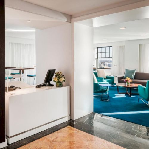 This image shows a modern, spacious office reception area with a check-in counter, seating in turquoise chairs, large windows, and decorative elements.