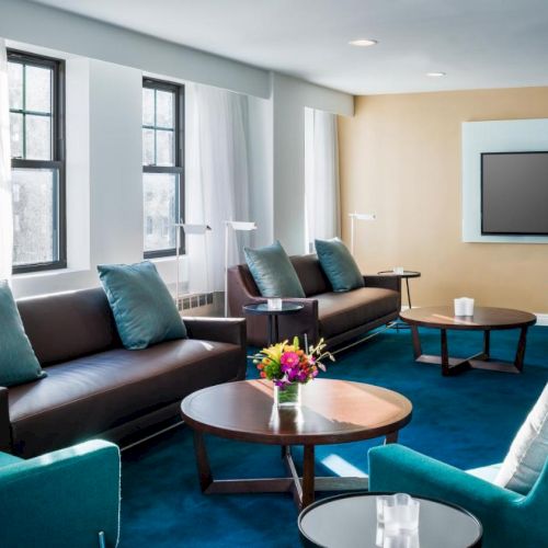 A modern living room with blue carpet, brown sofas, teal chairs, coffee tables, a mounted TV, and large windows with white curtains.