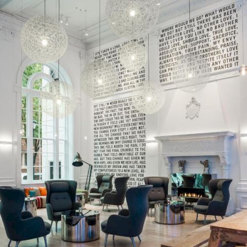 A modern room with high ceilings, artistic light fixtures, black chairs, a wooden bench, and abstract wall art. Large windows allow natural light inside.